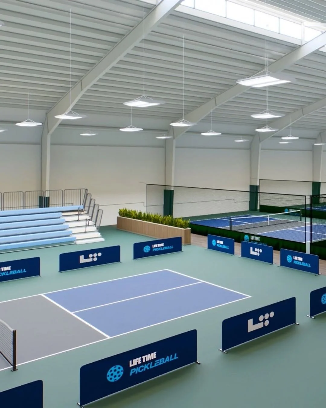 Indoor pickleball court with blue and gray surface, surrounded by blue barriers with 'LIFE TIME PICKLEBALL' and logo, and a seating area with bleachers and a planter with greenery.