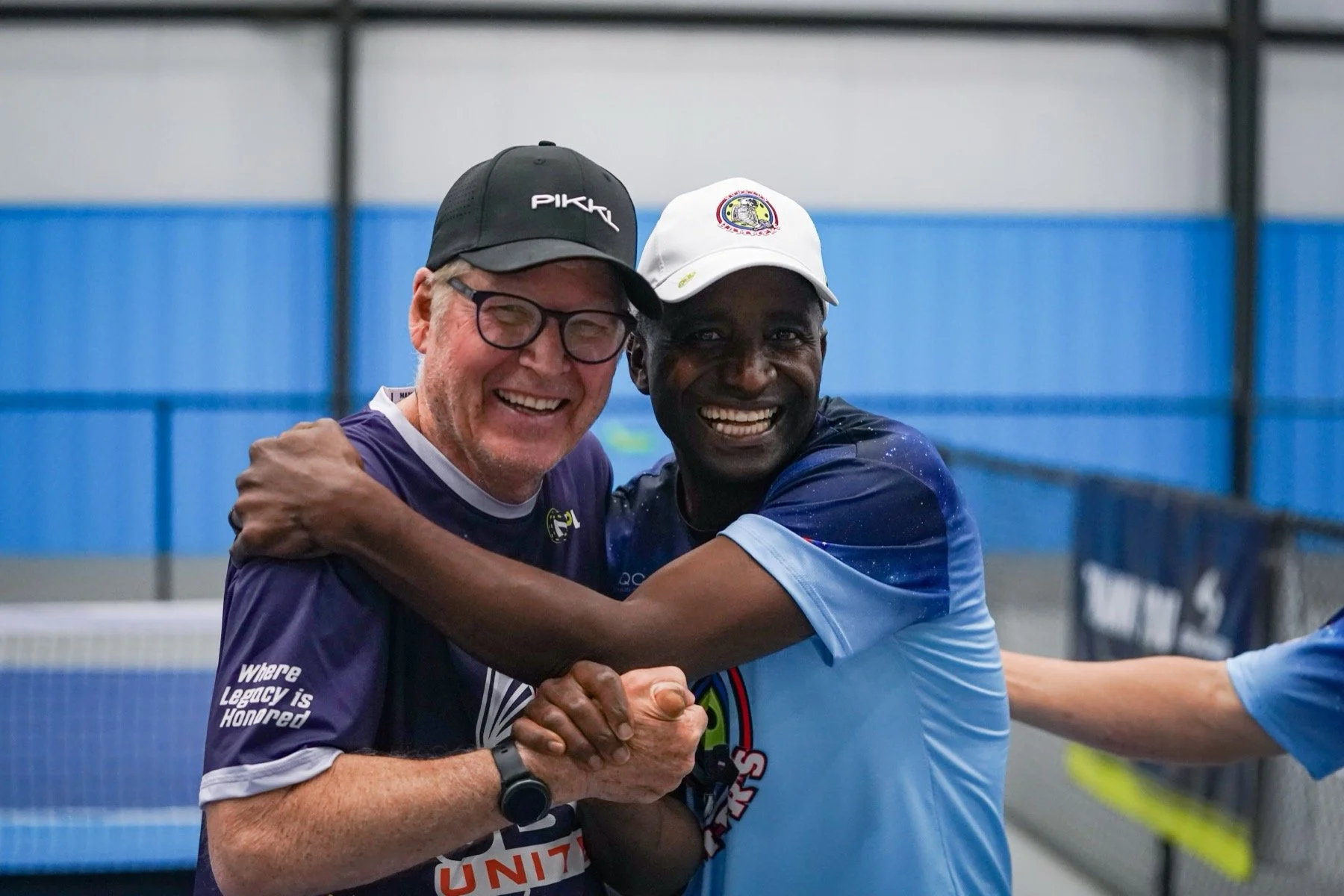 Two men smiling and embracing each other during a sports event in an indoor arena, wearing athletic clothing and hats, with a blue and white background.