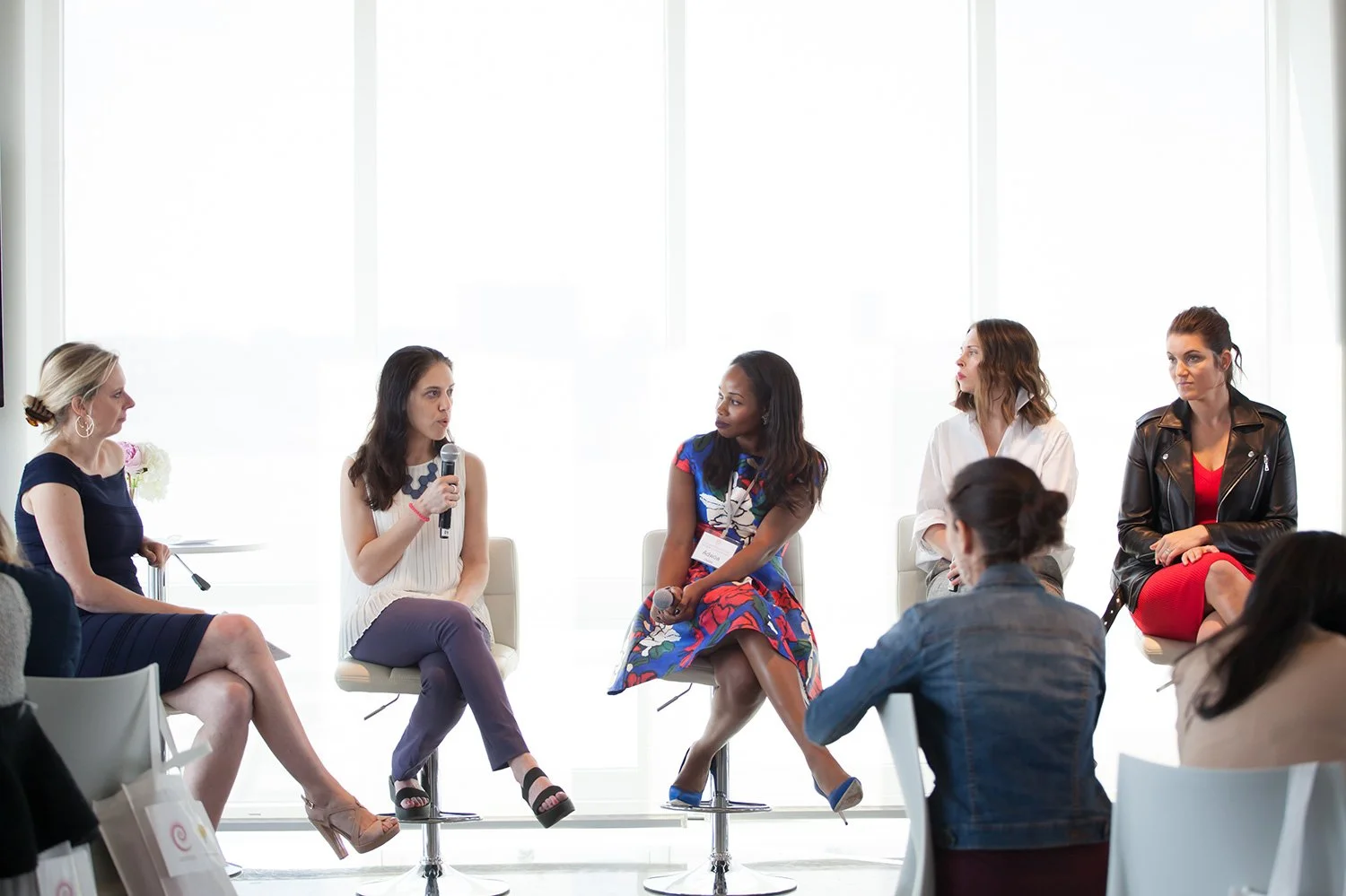 Women participating in panel discussion in a bright conference room, sitting on high chairs, with one woman speaking into a microphone.