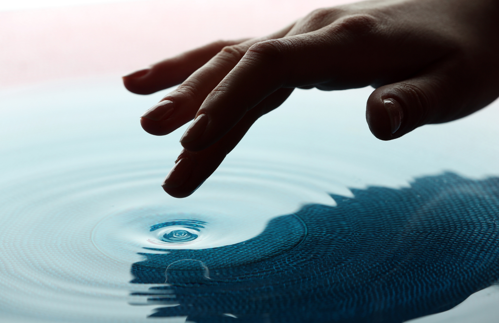 Close-up of a hand with fingers slightly bent touching the surface of water, creating ripples on the water's surface.