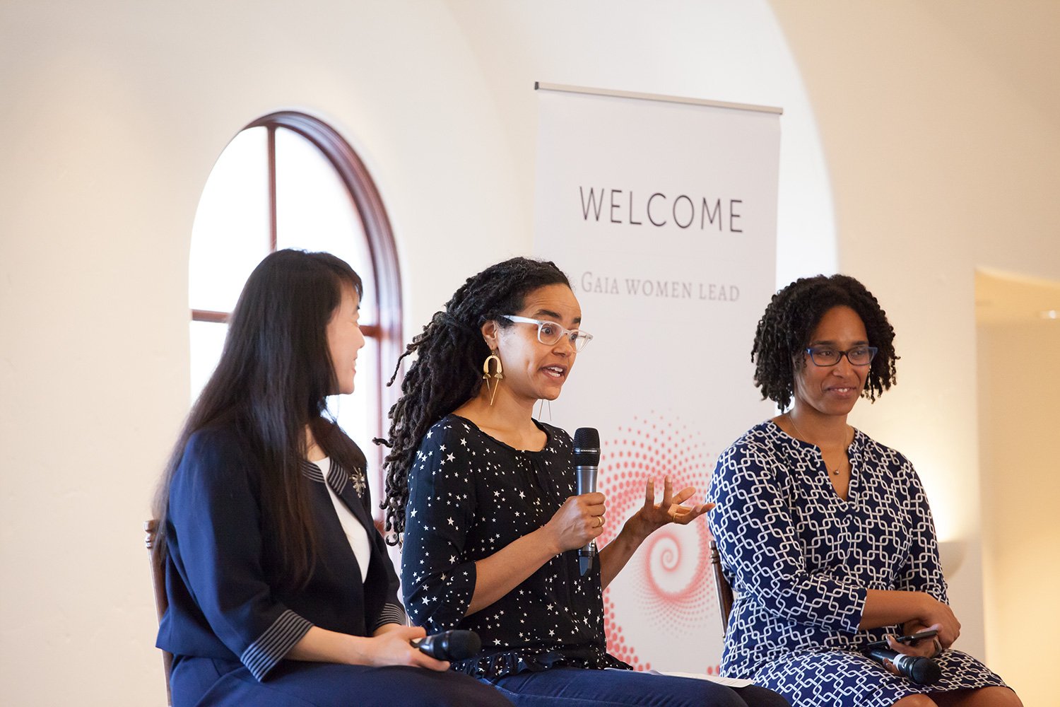 Three women seated on a panel, one speaking into a microphone, with a white banner behind them that reads "WELCOME GAIA WOMEN LEAD."