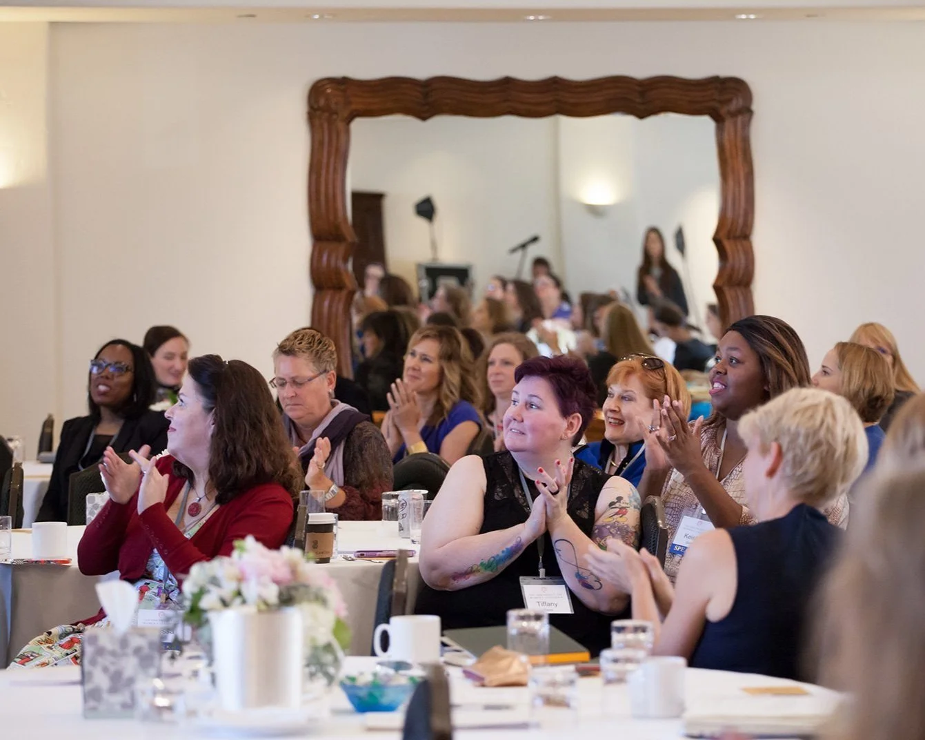 Women attending a conference or seminar, sitting at tables, clapping and smiling, with a large decorative mirror behind them reflecting the audience.