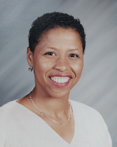 Rosalind Wilkes, a woman with short curly black hair smiling at the camera, wearing a white top and jewelry, against a gray background.
