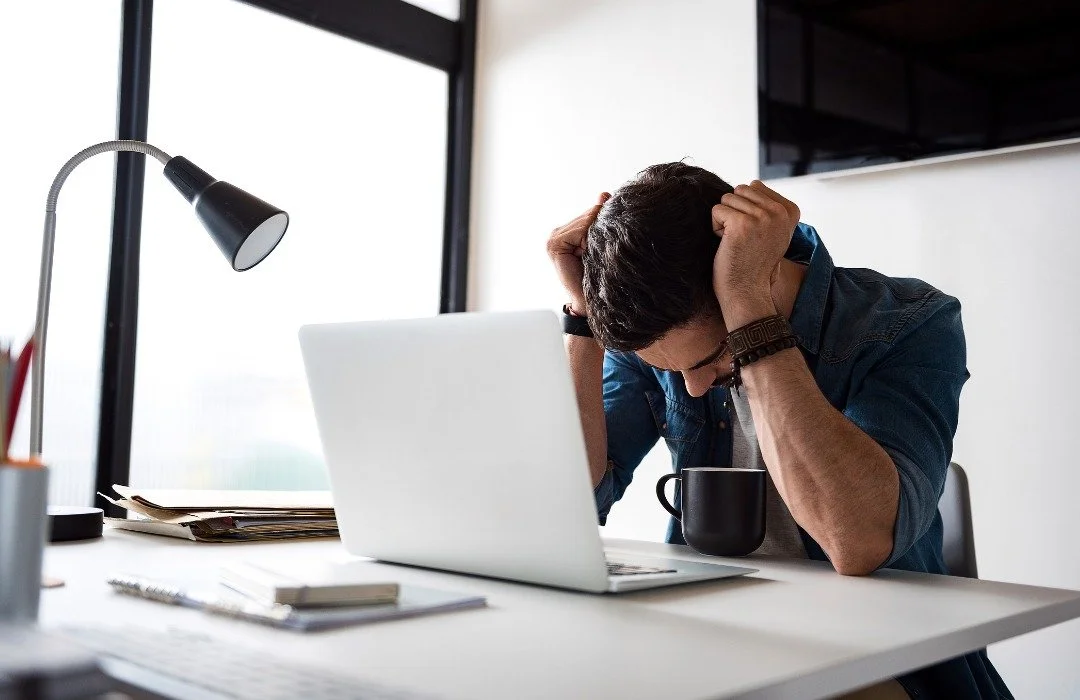 Young man sitting at a cluttered desk in an office, holding his head in stress, with a laptop, coffee mug, and papers.