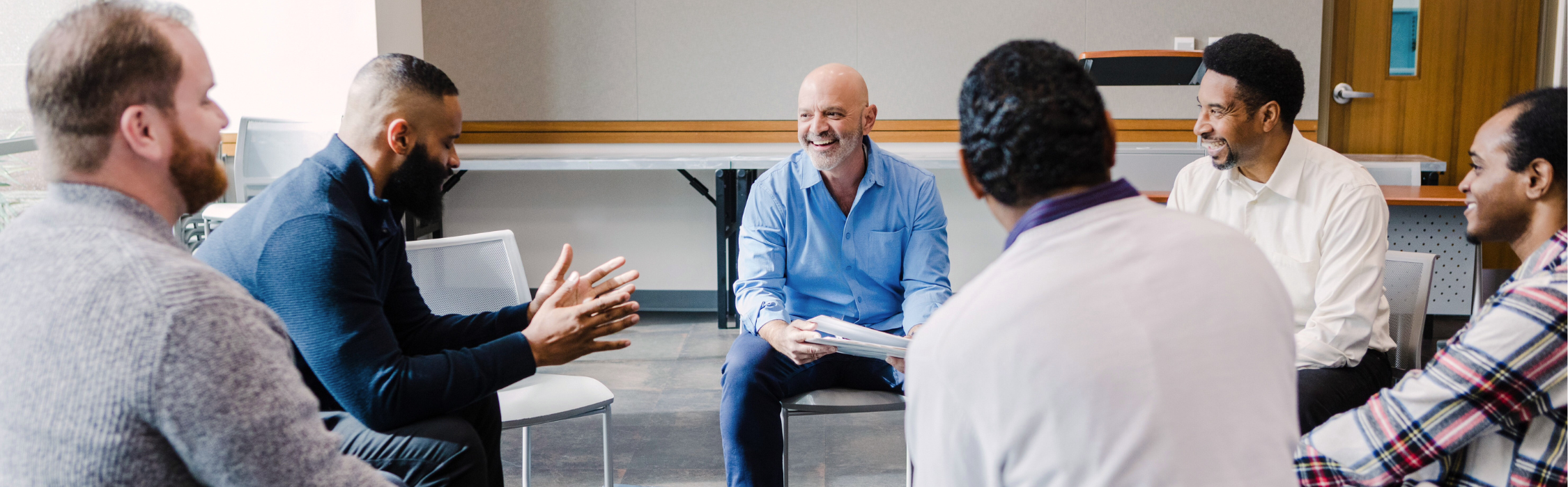 Group of diverse men sitting in a circle, engaged in conversation and smiling in a professional setting.