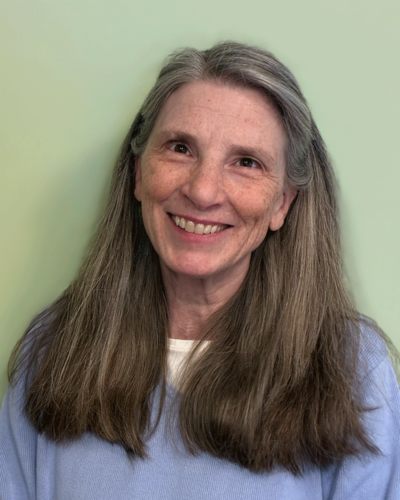Victoria Pekar, a smiling middle-aged woman with long gray hair, wearing a light blue top, standing against a light green background.
