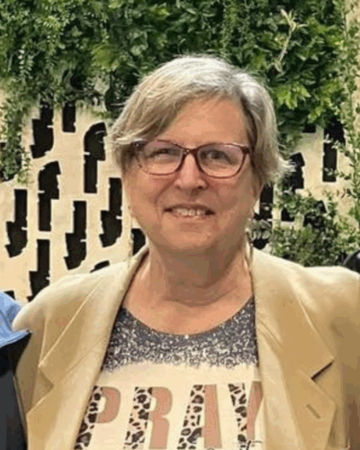A smiling woman with short gray hair and glasses wearing a beige blazer and a patterned shirt, standing outdoors with green foliage and a decorative fence in the background.