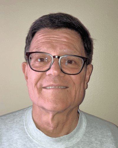 A middle-aged man with dark hair, glasses, and a gray shirt, smiling against a plain beige wall.