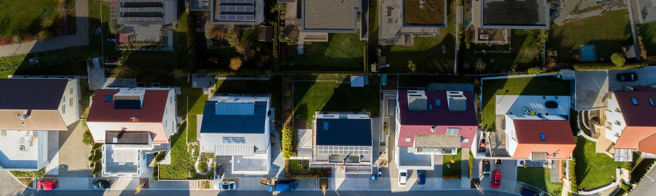 Aerial view of a residential neighborhood with several houses, green lawns, trees, and parked cars.