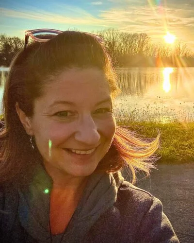 A woman smiling outdoors near a body of water during sunset, with trees in the background and sunglasses on her head.