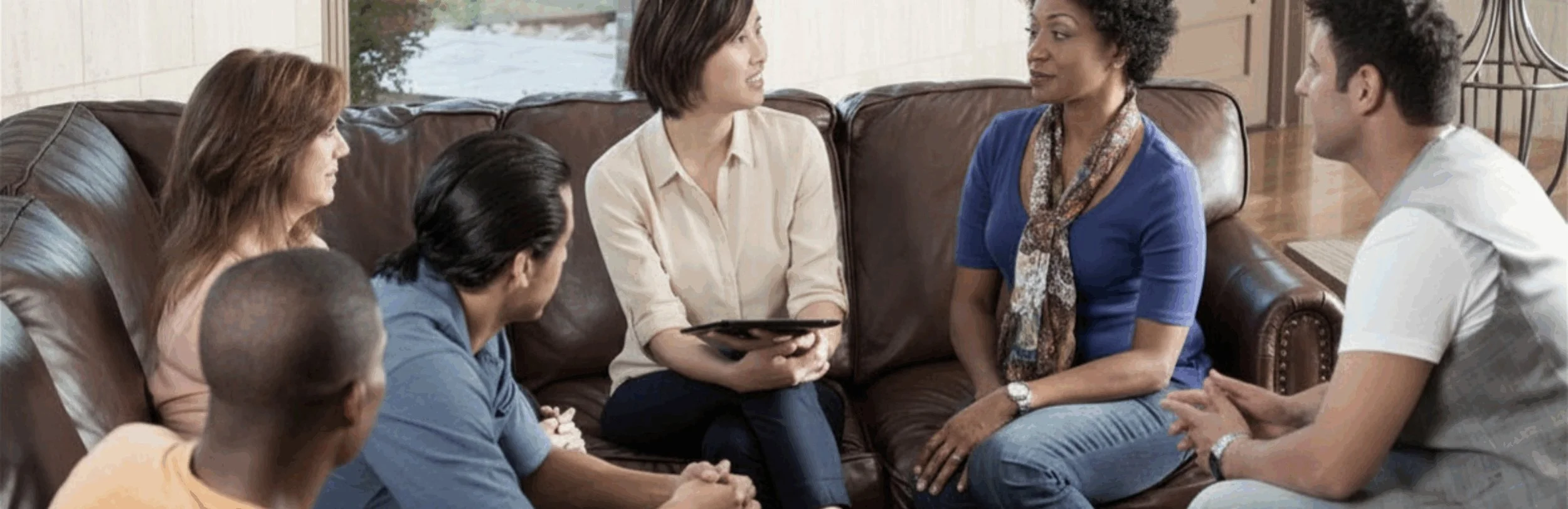 Group of six people sitting on a brown leather couch having a discussion. One woman is holding a tablet.
