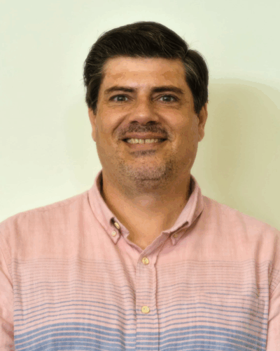 A man with dark hair and a slight beard smiling, wearing a pink button-up shirt, standing against a light-colored background.