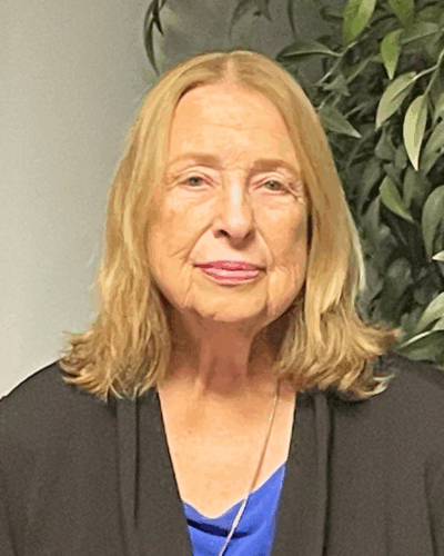 An older woman with shoulder-length blonde hair, wearing a black jacket over a blue top, standing indoors in front of a green leafy plant.