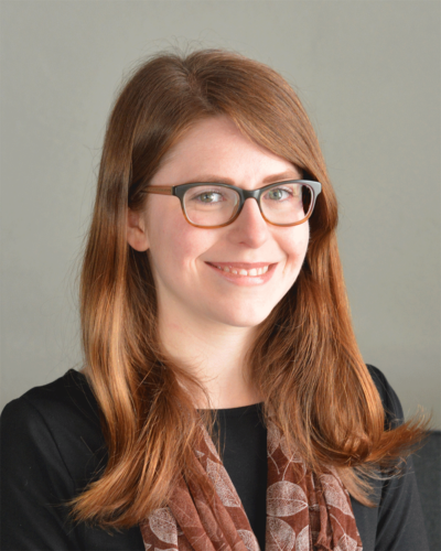 Grace Rybak, a woman with red hair and glasses smiling at the camera, wearing a black top and a patterned scarf, against a gray background.