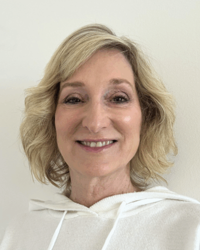 A smiling woman with shoulder-length blonde hair, wearing a white hoodie, standing against a plain off-white wall.