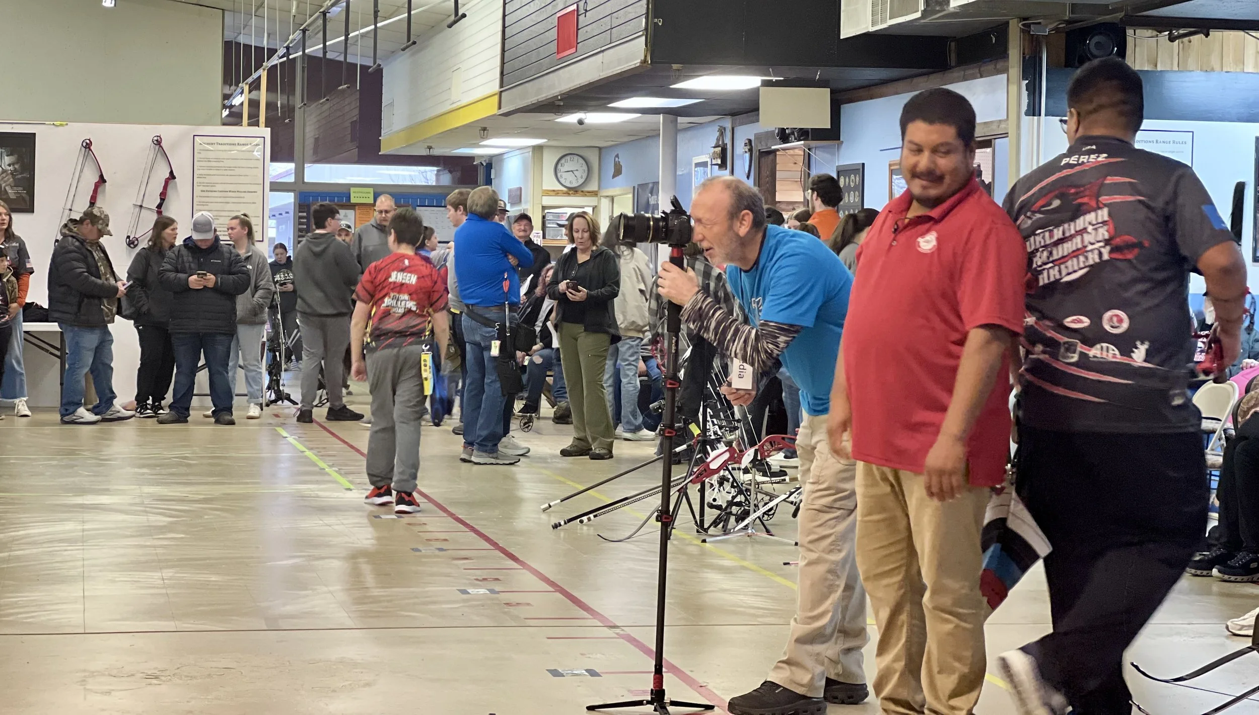 People gathering at indoor archery event, some preparing or aiming bows, others observing, with equipment and posters on walls.