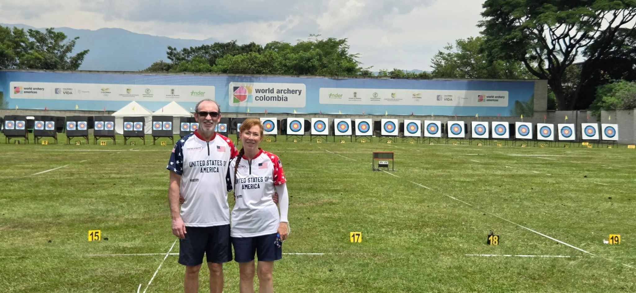 Two archers standing in front of an archery target range at the World Archery Americas event in Colombia, dressed in United States of America team uniforms, with targets and a banner in the background.