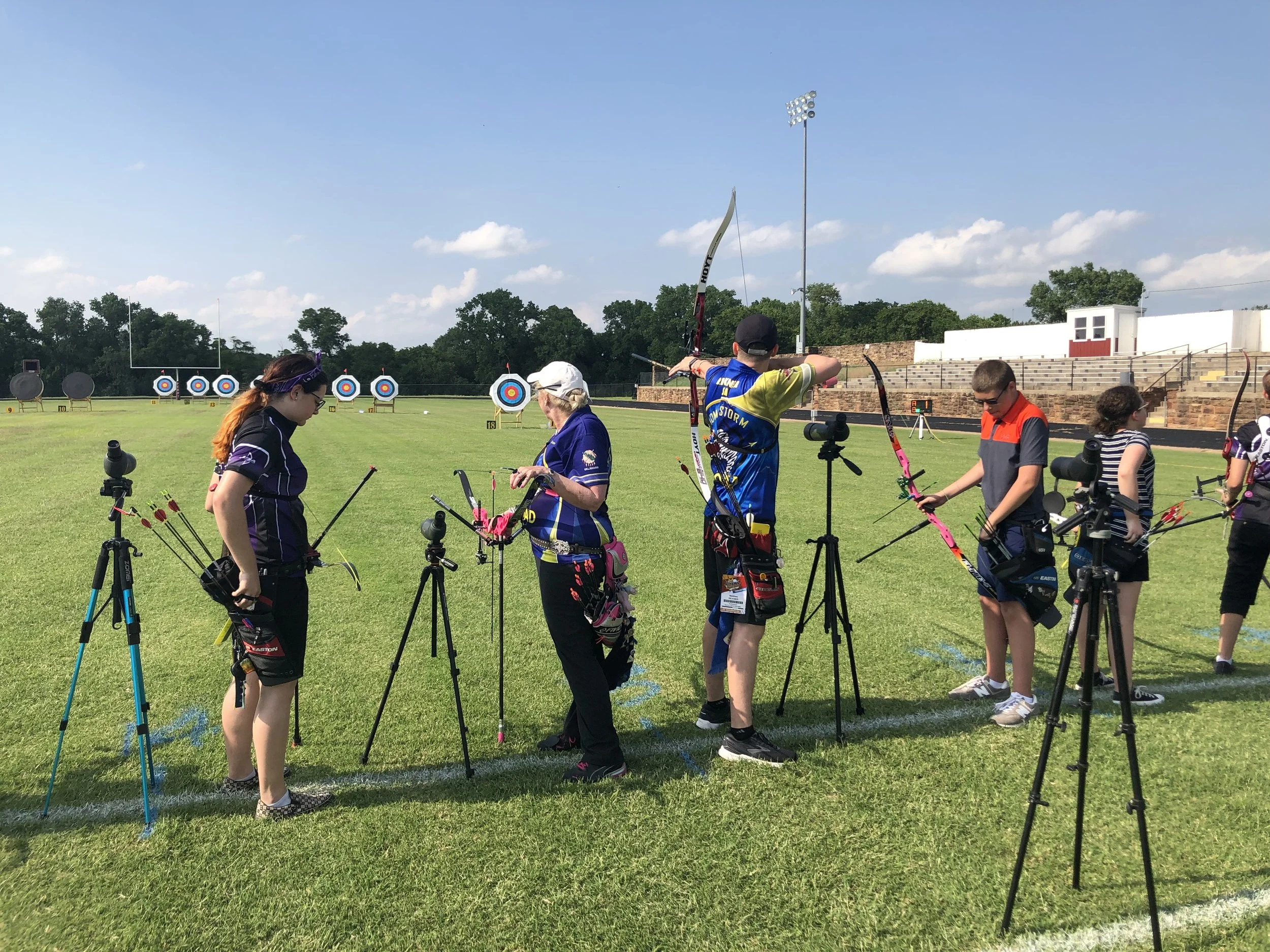 Group of people participating in archery practice on a grassy field with targets in the background, under a clear blue sky.