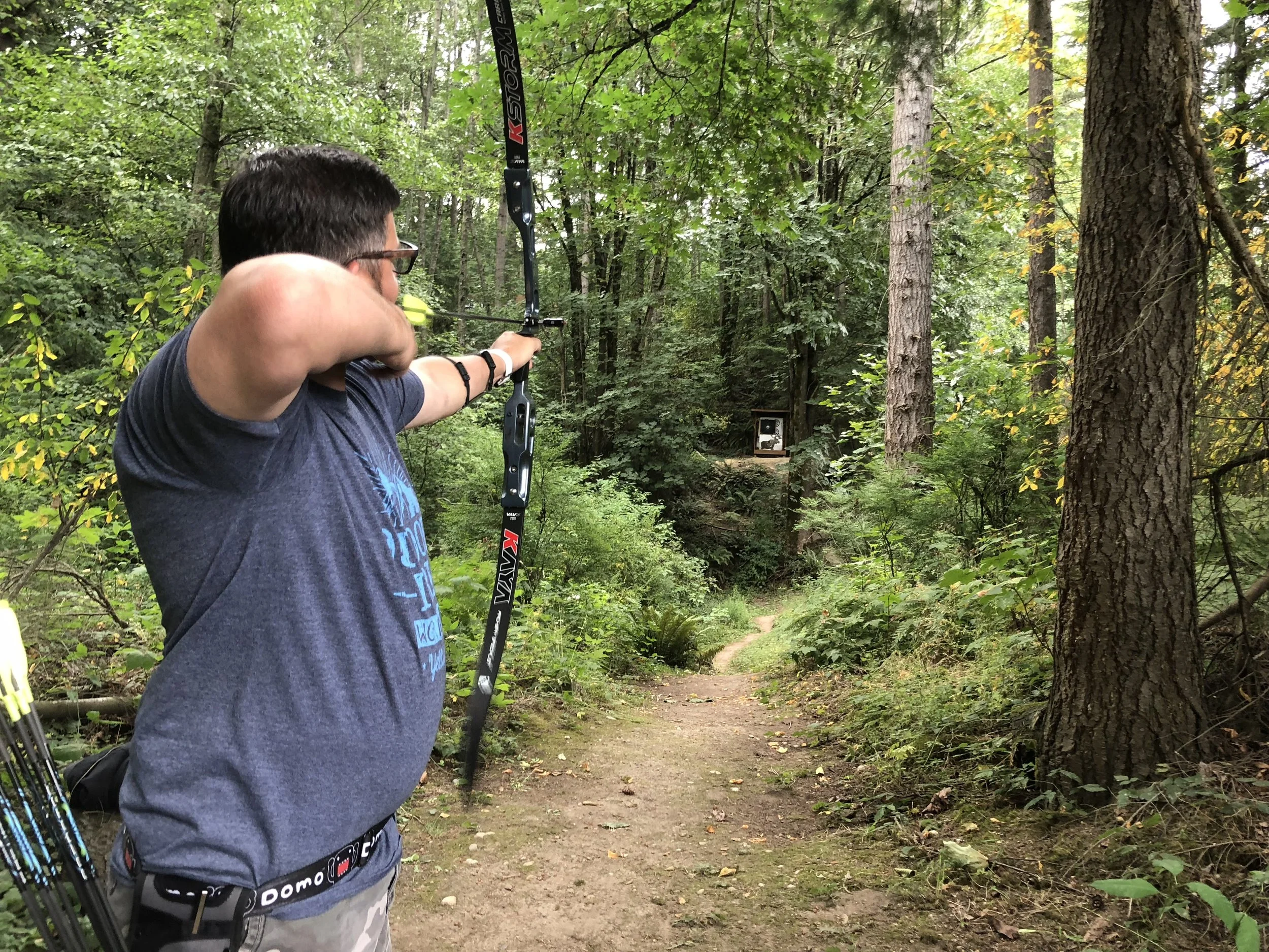 An archer preparing to shoot in a green wooded field course, aiming a black bow at a target down a dirt trail surrounded by tall trees and lush foliage.