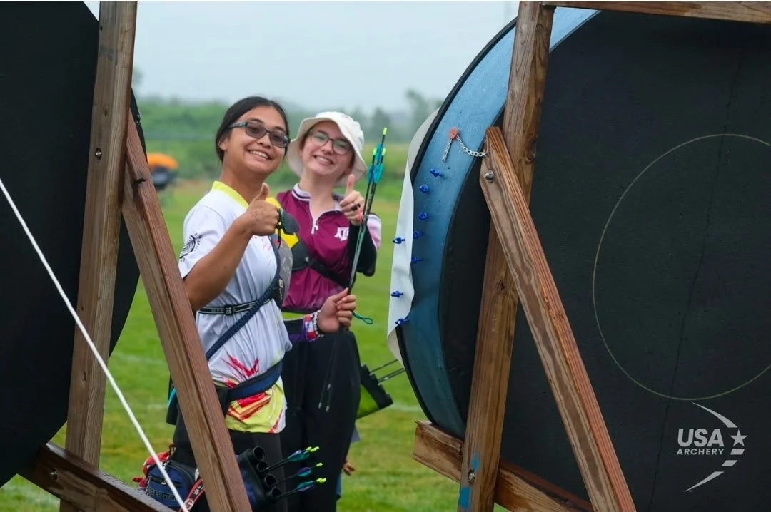 Two archers smiling and giving a thumbs-up at an outdoor archery range, holding bows and arrows, standing next to targets, with a cloudy sky and grassy field in the background.