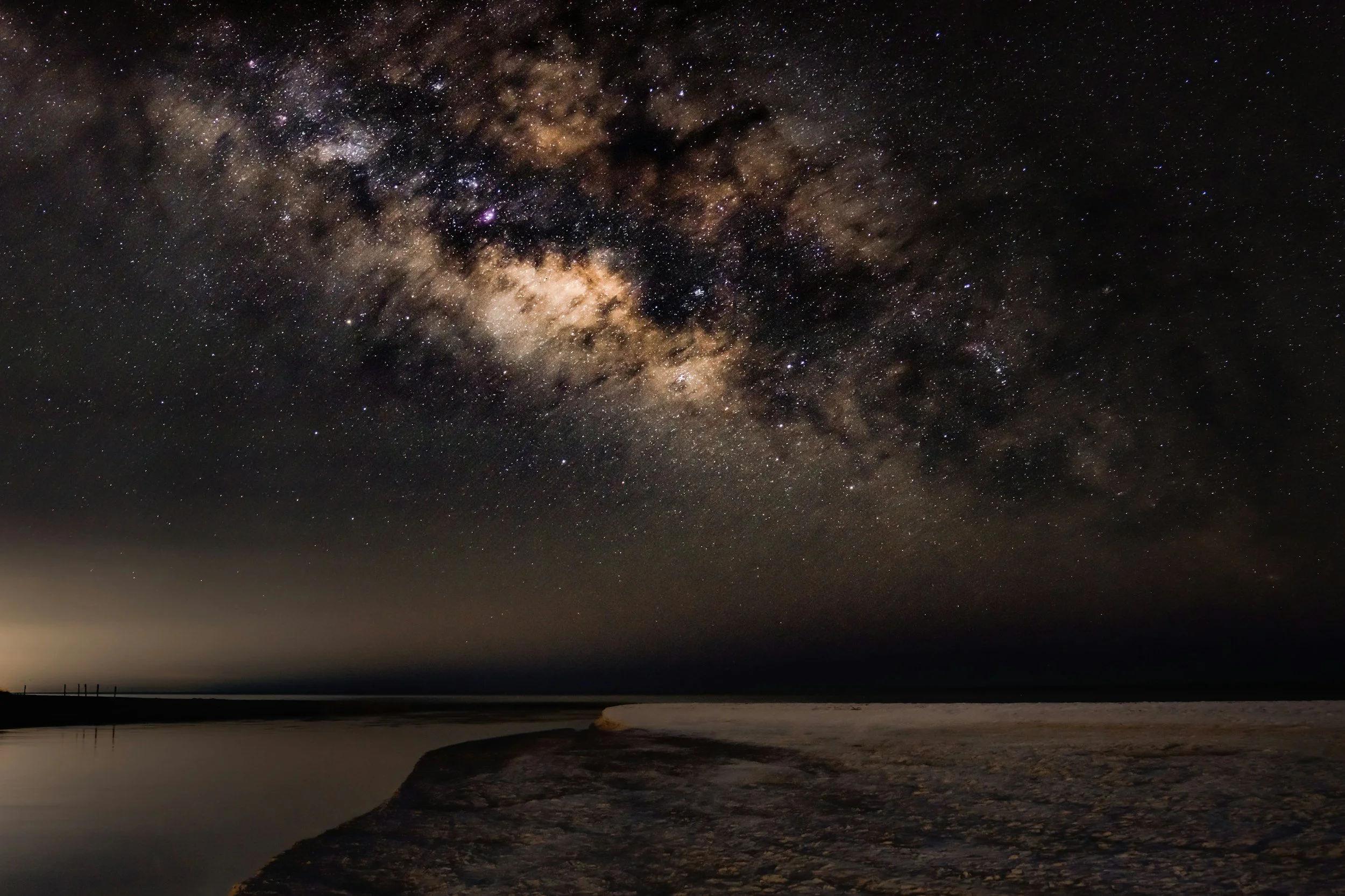 Night sky filled with stars and the Milky Way galaxy over a body of water and shoreline, with a silhouette of a distant pier.