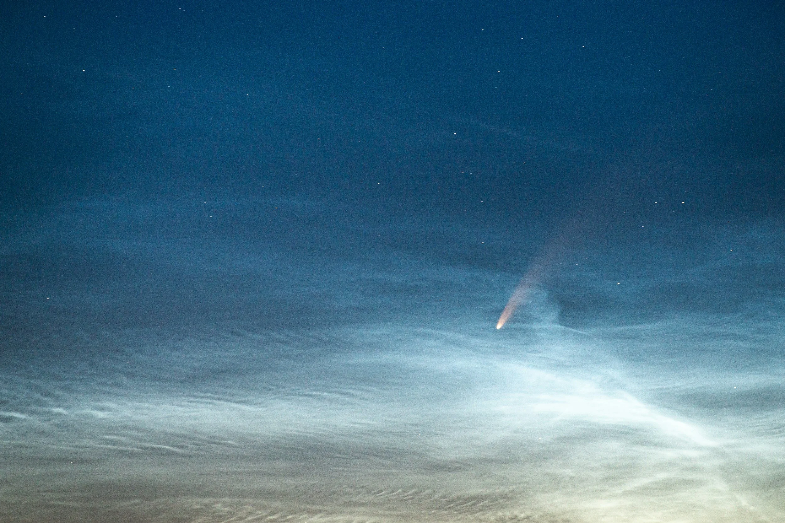 A photograph of the night sky above a body of water, showing a bright comet with a tail streaking across the sky.