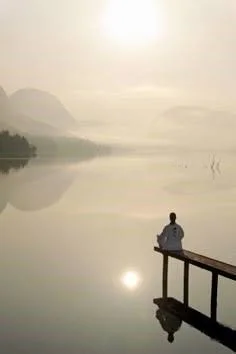 Person sitting on a dock overlooking a calm lake with mountains in the background, during sunrise or sunset.