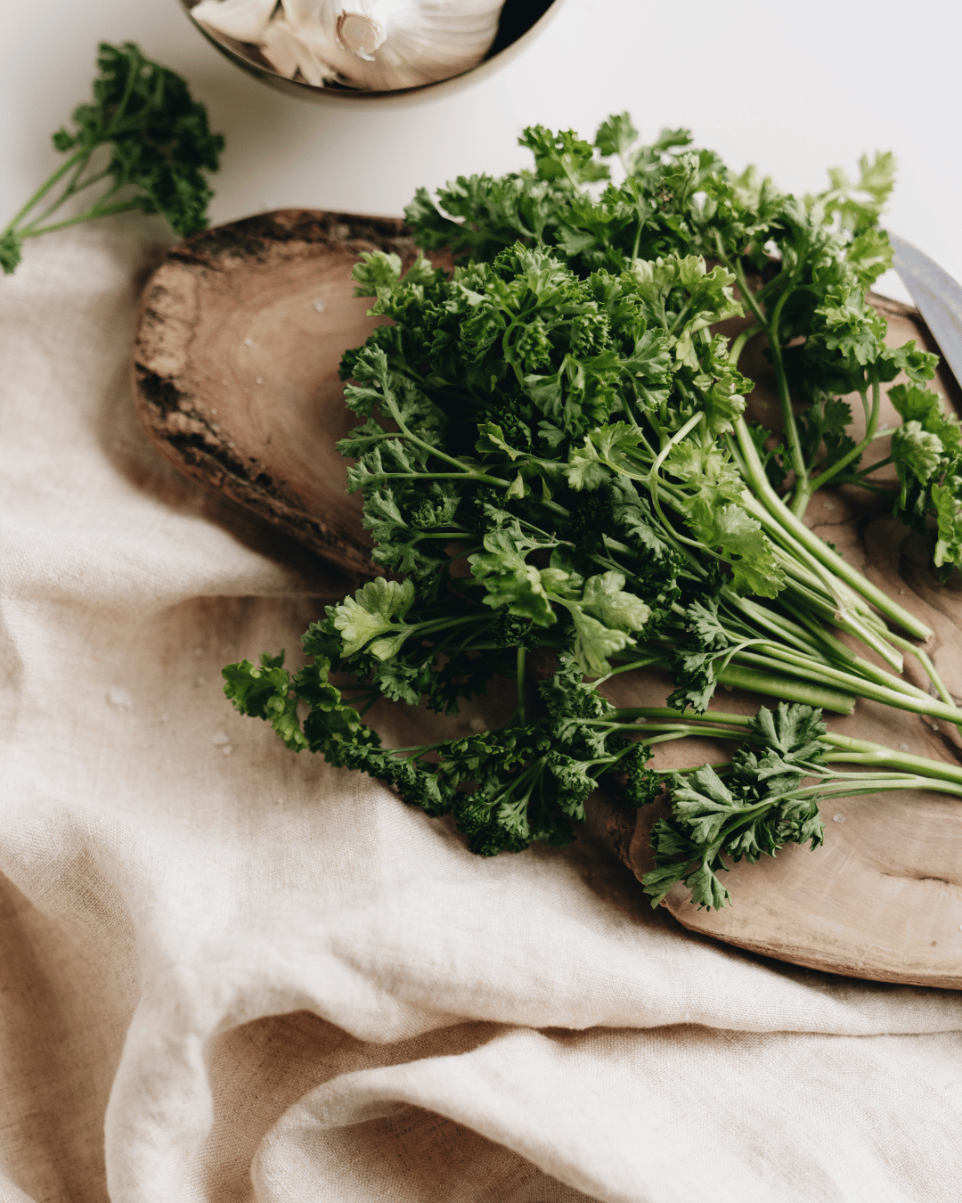 Fresh parsley bunch on a wooden cutting board with a knife nearby, on a cream-colored cloth surface.