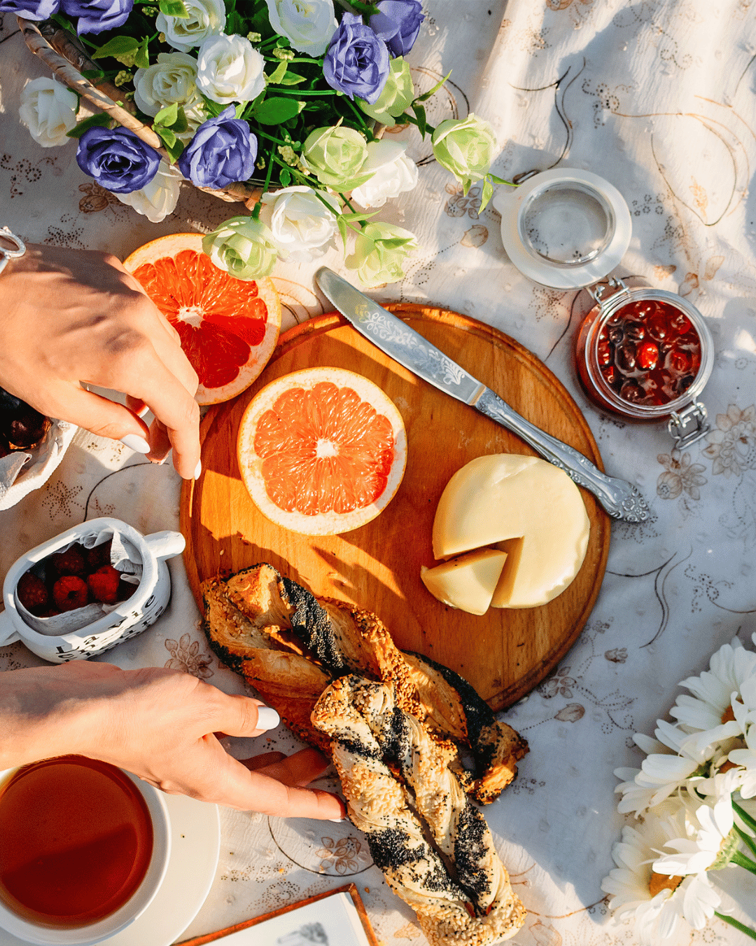 A picnic table set with sliced grapefruit, cheese, grilled fish, and a cup of tea, surrounded by a floral arrangement and jars of berries and cherries.