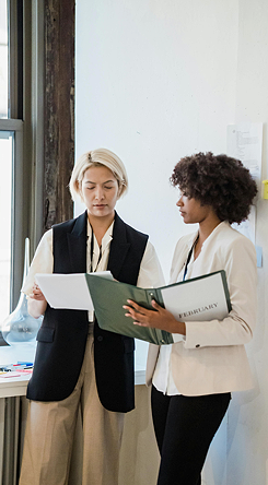 Two women standing indoors, looking at a binder together. One woman has blonde hair and is wearing a black vest and beige pants, while the other woman has curly hair and is wearing a light-colored blazer.