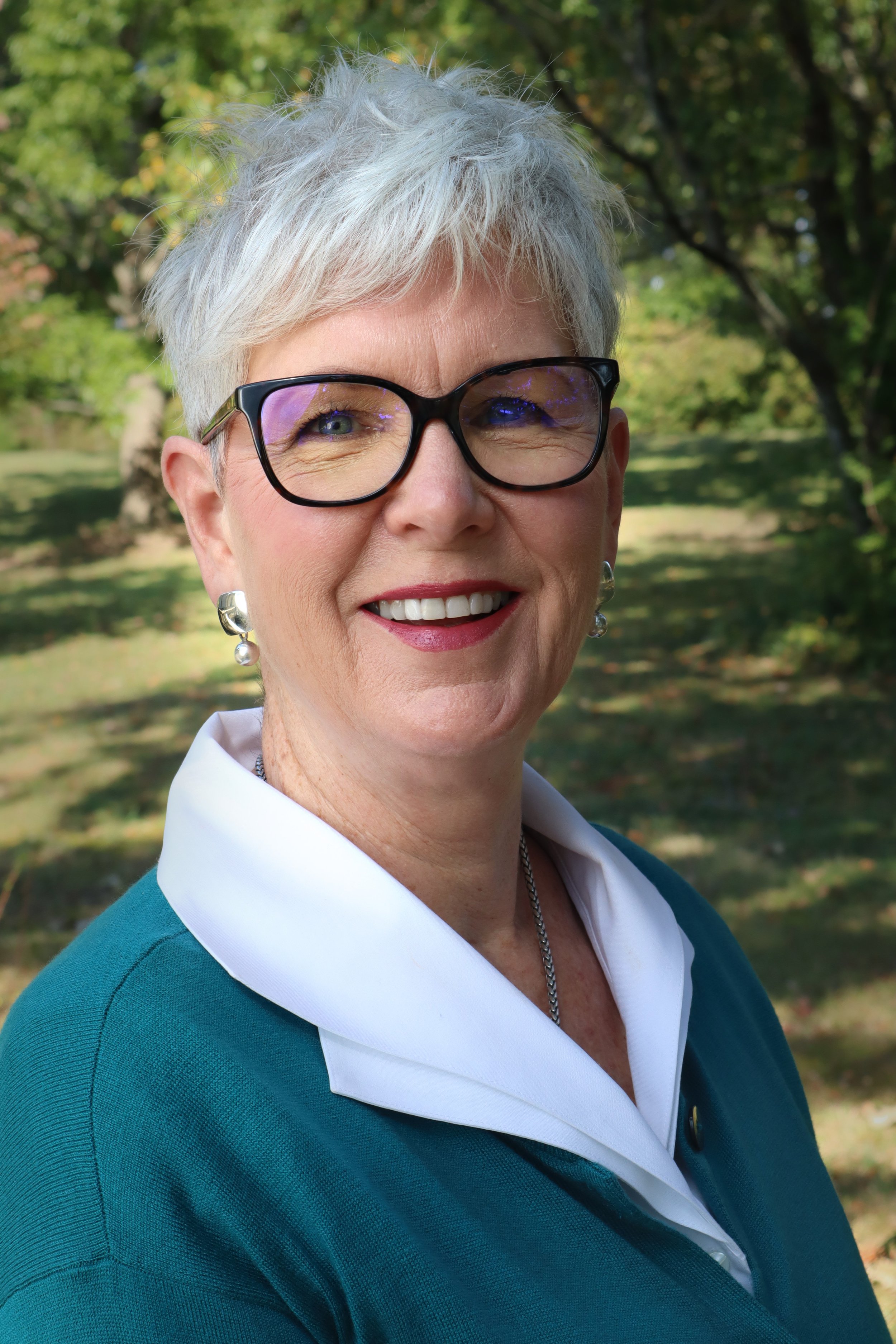 Close-up portrait of a smiling older woman with short gray hair, wearing glasses, pearl earrings, a white collared shirt, and a teal sweater, outdoors with green trees in the background.