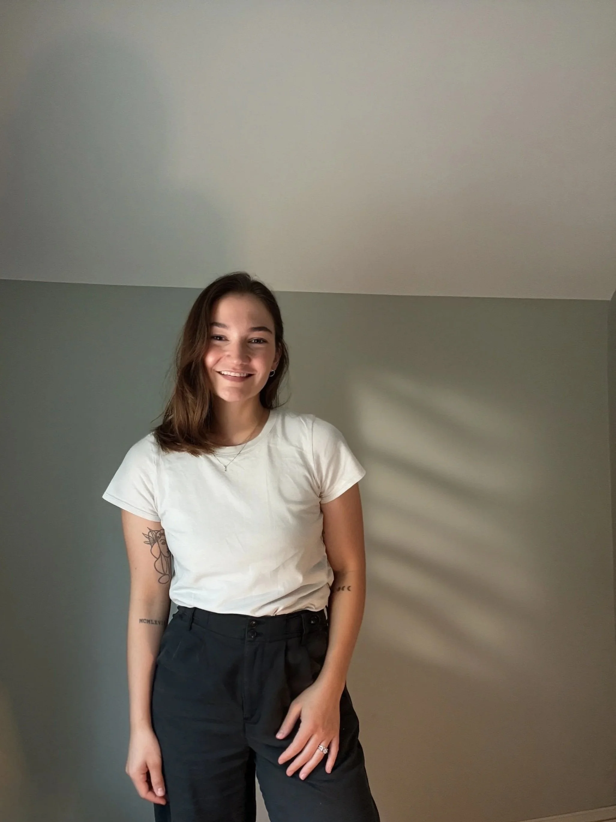 Photo of therapist Selena Lindamood, woman with brown hair smiling, wearing a white t-shirt and black pants, standing against a gray wall