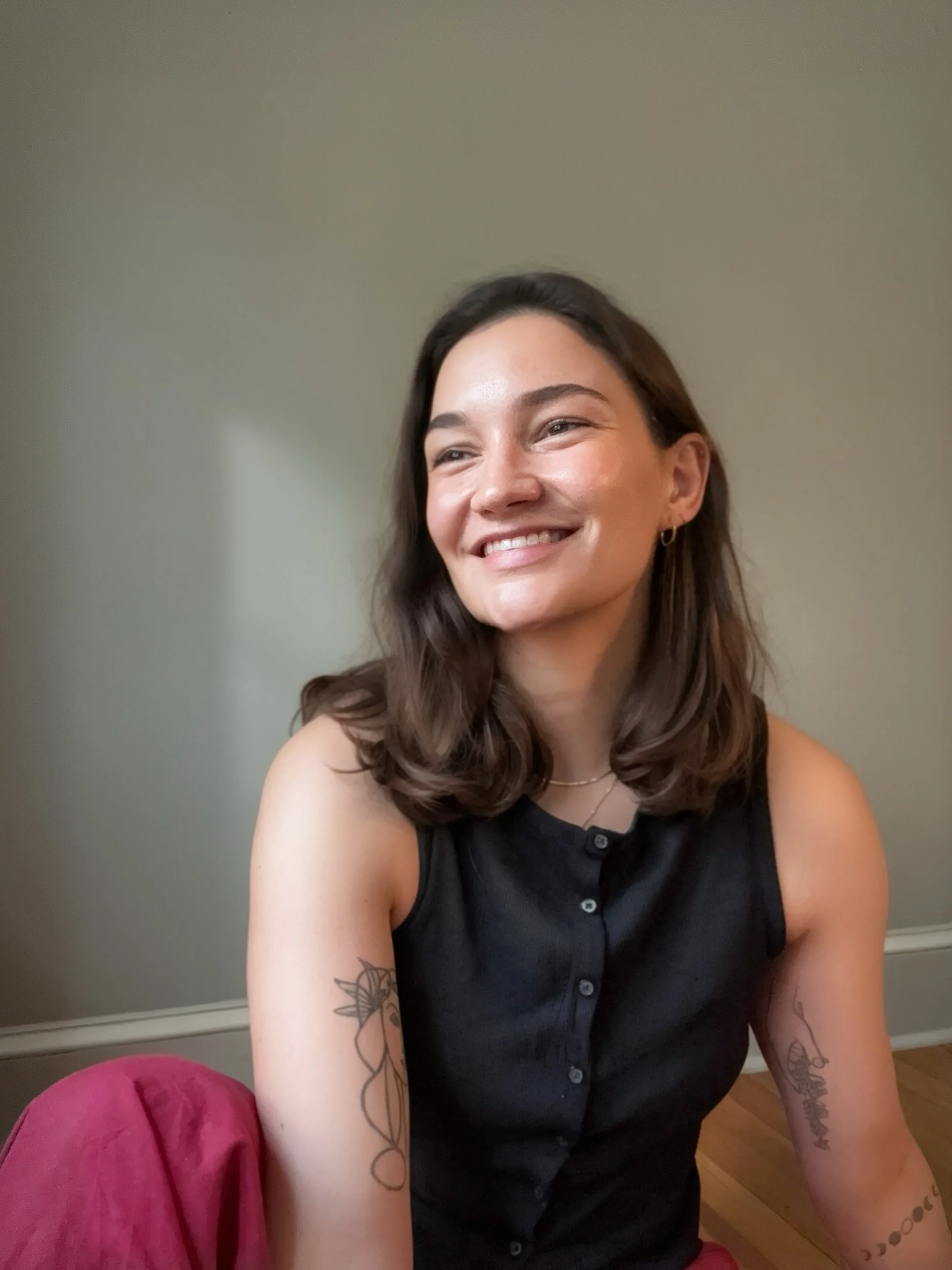 Therapist Selena Lindamood LPC, a woman smiling, sitting on a pink couch in a room with a light-colored wall and wooden floor, wearing a black sleeveless button-up top, with tattoos on her arms and ear piercings.