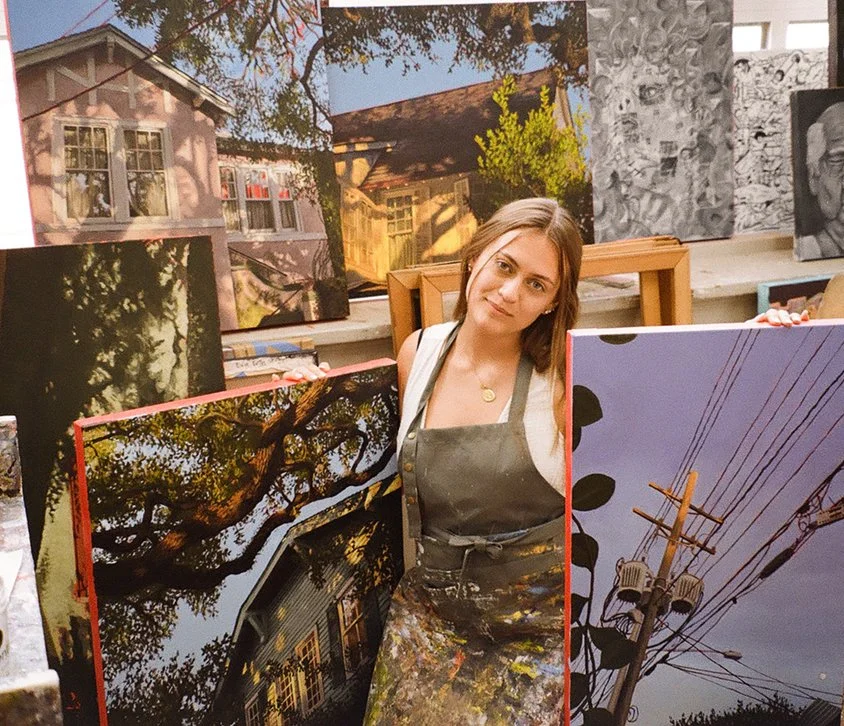 Evie Roth in a painting apron stands in her college art studio, holding up her paintings of trees and houses, with additional artwork of trees and power lines in the background.