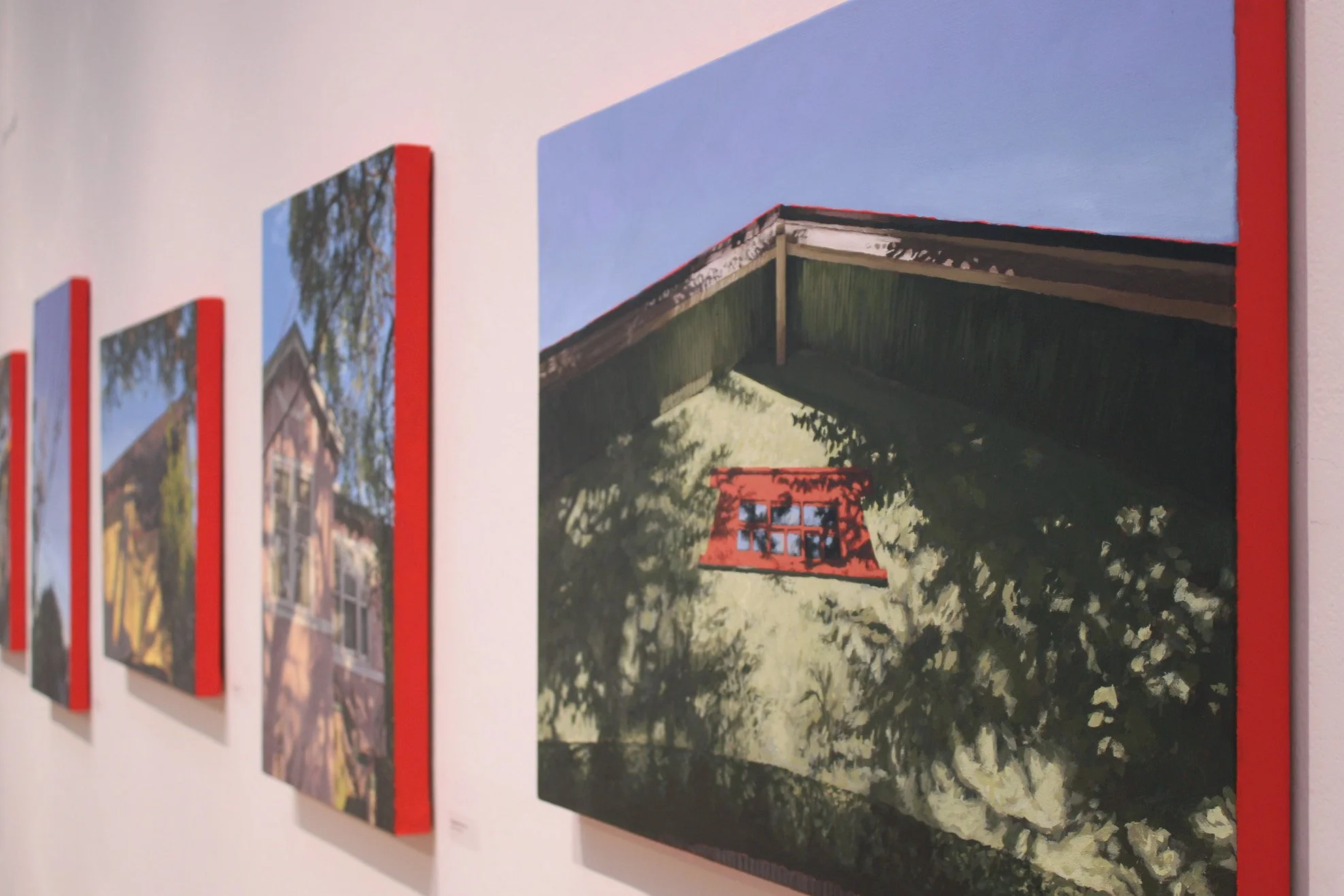 A series of landscape paintings of houses and trees displayed on a white gallery wall, each with a red border on the side.