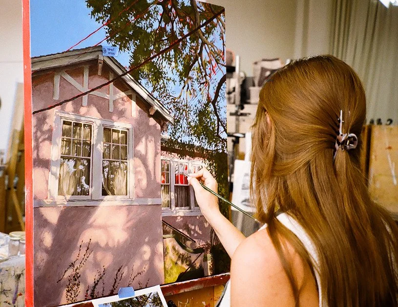 A woman with long red hair, wearing a white sleeveless top, is painting a mural of a house on a canvas. The house in the mural is pink with white trim and has multiple windows. The woman is standing indoors in an art studio, surrounded by art supplies and equipment.
