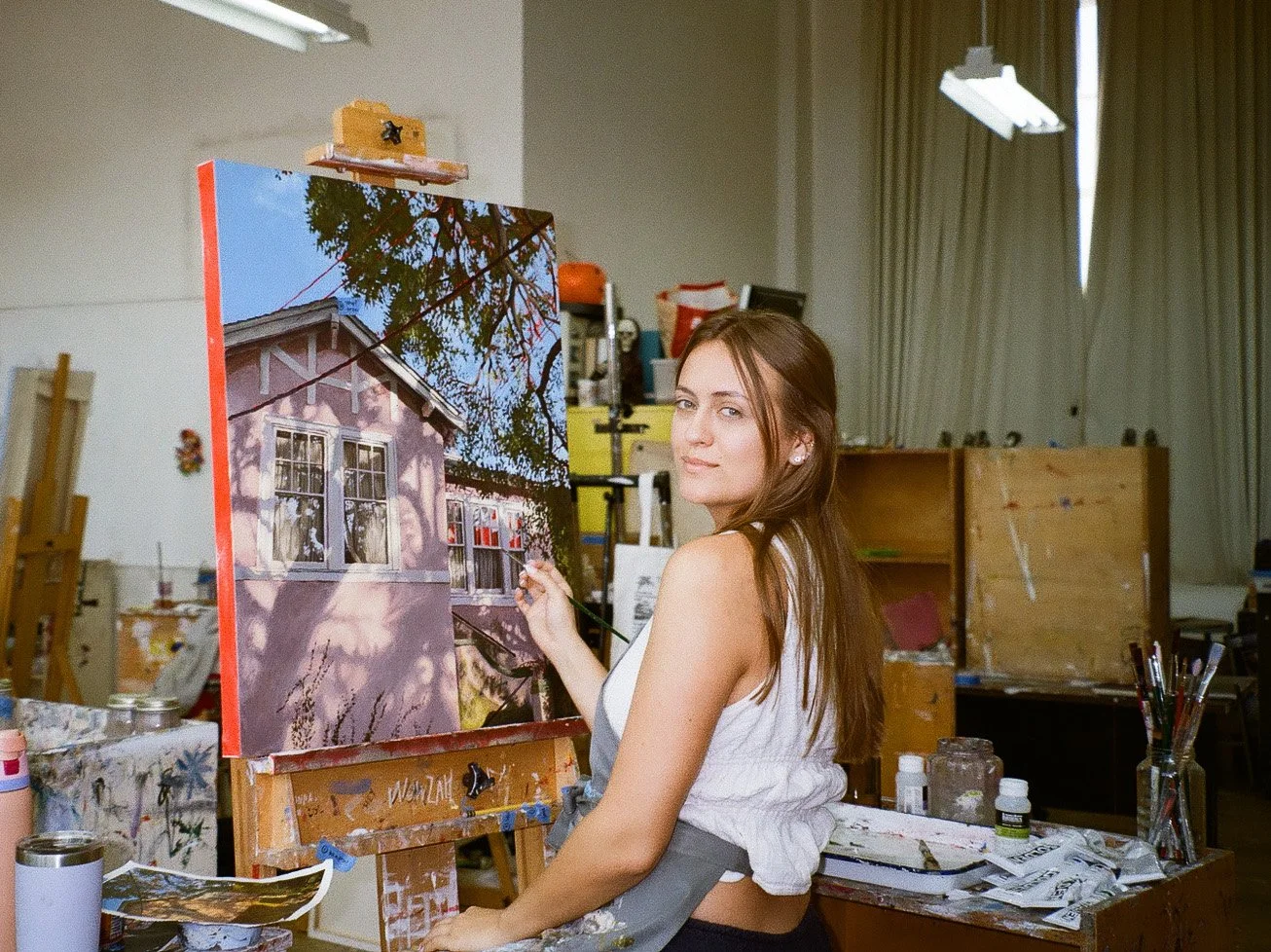 Evie Roth in a painting apron stands in her college art studio, painting a house on a canvas mounted on an easel surrounded by art supplies and clutter.