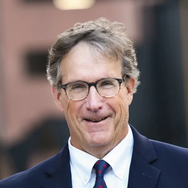 Portrait of a smiling gray-haired man wearing black glasses, a navy suit, white shirt, and red-blue tie in a softly blurred setting.