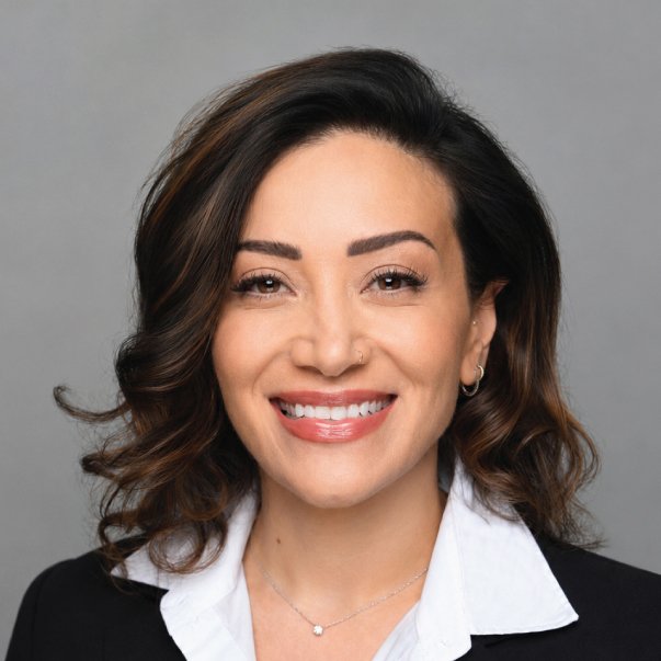 Professional headshot of a smiling woman with wavy dark hair in a black blazer and white blouse against gray background.