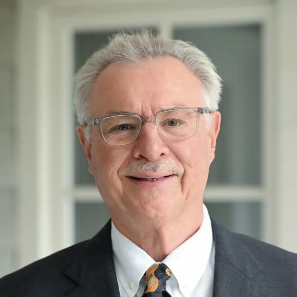Professional headshot of a smiling older man with gray hair and glasses, wearing a dark suit and patterned tie before bright windows.