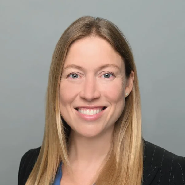 Professional headshot of a smiling woman with long blonde hair in a black blazer against a neutral background.