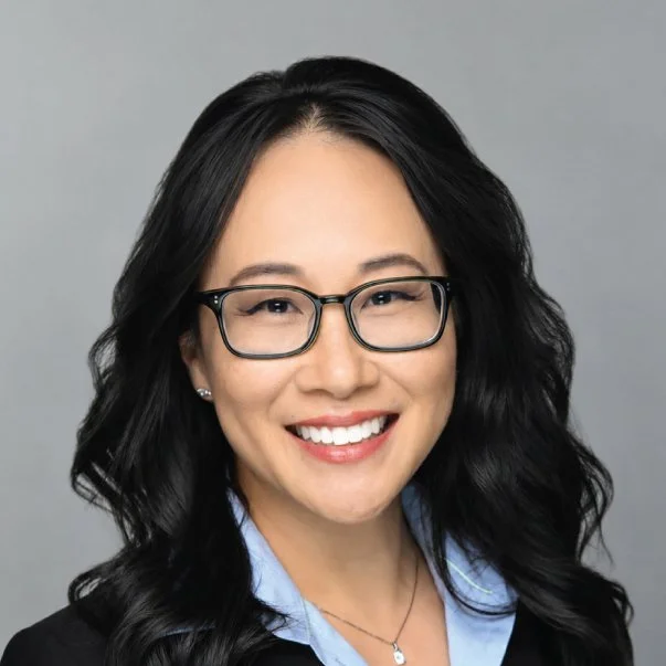 Professional headshot of a smiling woman with long black hair and glasses, wearing a blue collared shirt against a gray background.