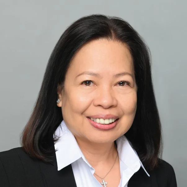 Professional headshot of a smiling woman with shoulder-length dark hair, wearing a black blazer, white collar, and silver necklace on gray background.