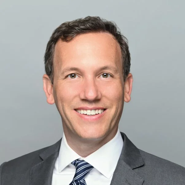 Professional headshot of a smiling man in a gray suit, white shirt, and striped tie against a neutral gray background.