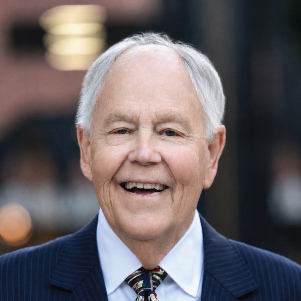Close-up portrait of an older white-haired man smiling warmly in a navy pinstripe suit and patterned tie, with a softly blurred background.