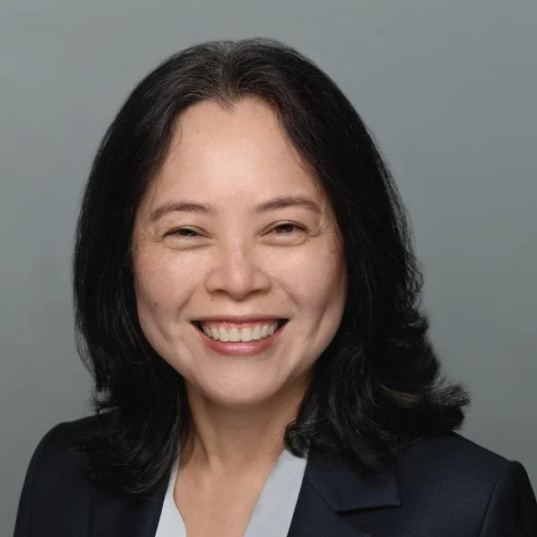 Professional headshot of a smiling woman with shoulder-length black hair, wearing a dark blazer and gray blouse against a gray background.