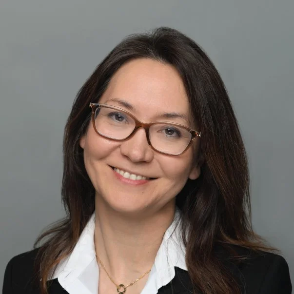 Professional headshot of a smiling woman with long dark hair and glasses, wearing a white shirt and black blazer against a gray background.