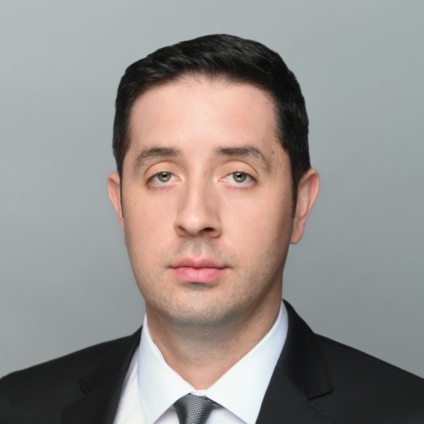 Professional headshot of a serious-looking man with short dark hair wearing a black suit and white shirt against a gray background.