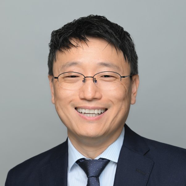 Professional headshot of a smiling man wearing glasses, a navy suit, and tie against a gray background.