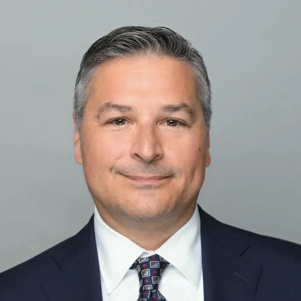 Professional headshot of man with salt-and-pepper hair in a navy suit and patterned tie, smiling calmly against a gray background.